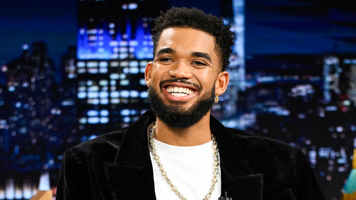 Karl-Anthony Towns smiles broadly on the set of The Tonight Show, dressed in a black velvet blazer over a white tee with a gold chain, celebrating his two championship rings during a guest appearance on the late-night program.