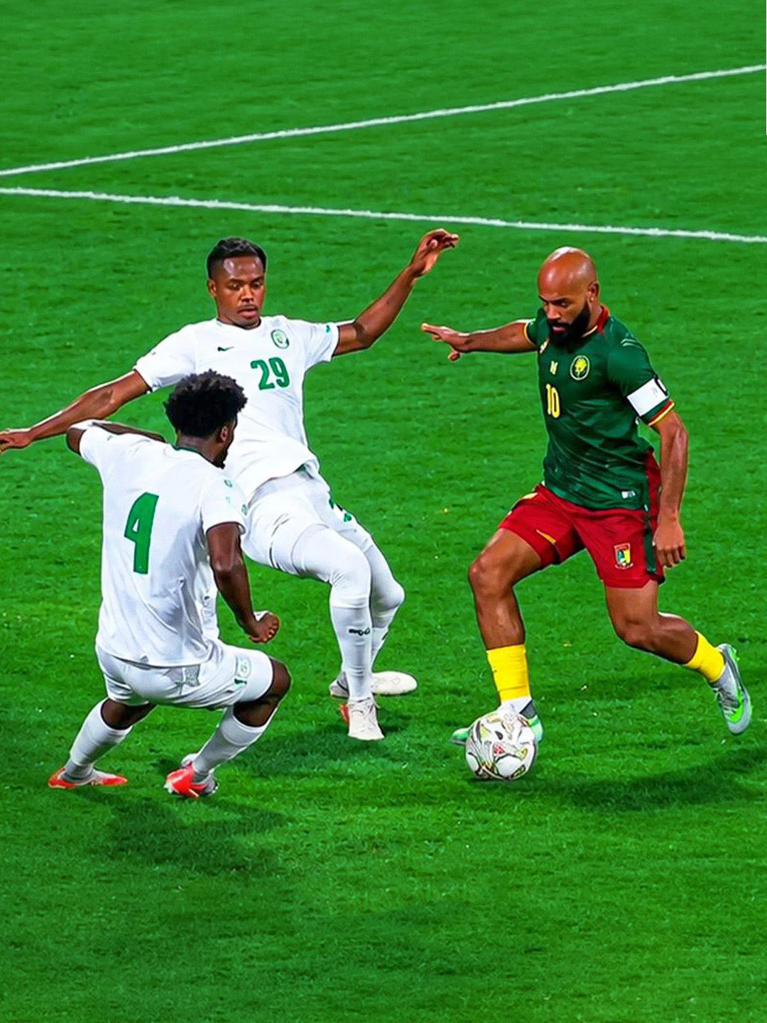 Bryan Mbeumo in a green, red and yellow Cameroon kit prepares to kick the ball past two Comoros players wearing all-white kits.