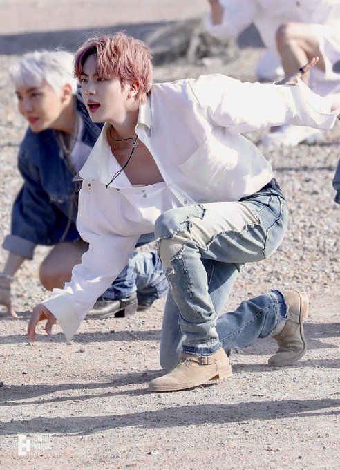 A performer with pink hair wearing a white shirt and distressed jeans crouches on a gravelly surface, with other dancers visible in the background.