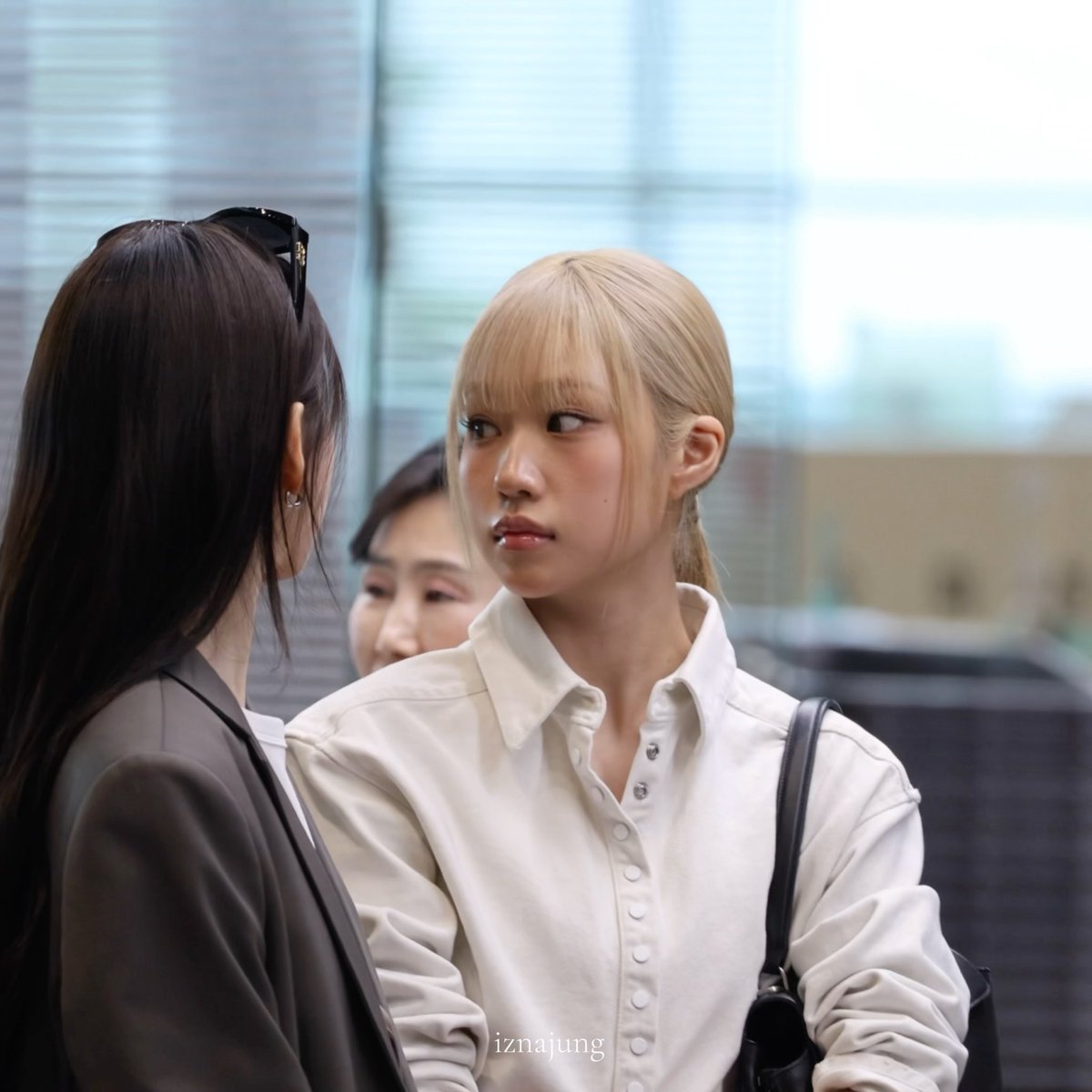 A blonde woman with bangs in a cream-colored button-up shirt looks sideways with a serious expression while standing near another woman with dark hair, watermarked 'iznajung'.