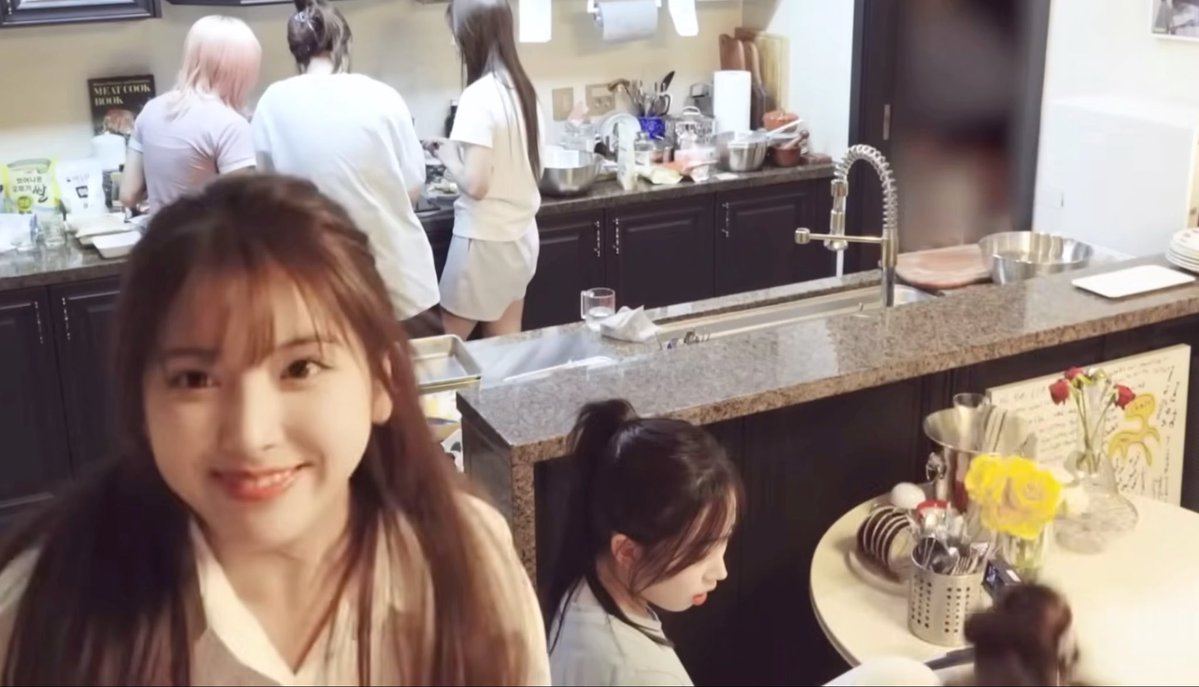 A selfie taken in a kitchen where a smiling woman is in the foreground, with other people working at the counter behind her, and a "MEAT COOK BOOK" sign visible on the wall.