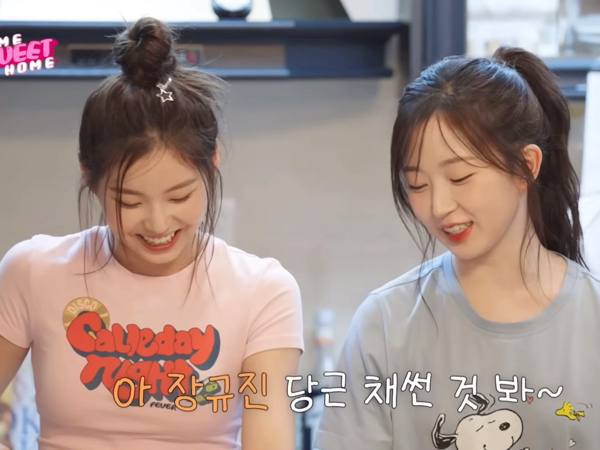 Two young women are smiling and looking down; the one on the left wears a pink 'Calyday Night Fever' t-shirt with a top knot, and the one on the right wears a light blue Snoopy t-shirt with a ponytail. Korean text overlay reads '아장꾸진 당근 채썬 것 봐~'.