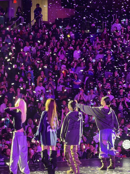 Four performers stand on a stage facing away from the camera, looking out at a large crowd holding up phones and confetti falling in purple lighting.