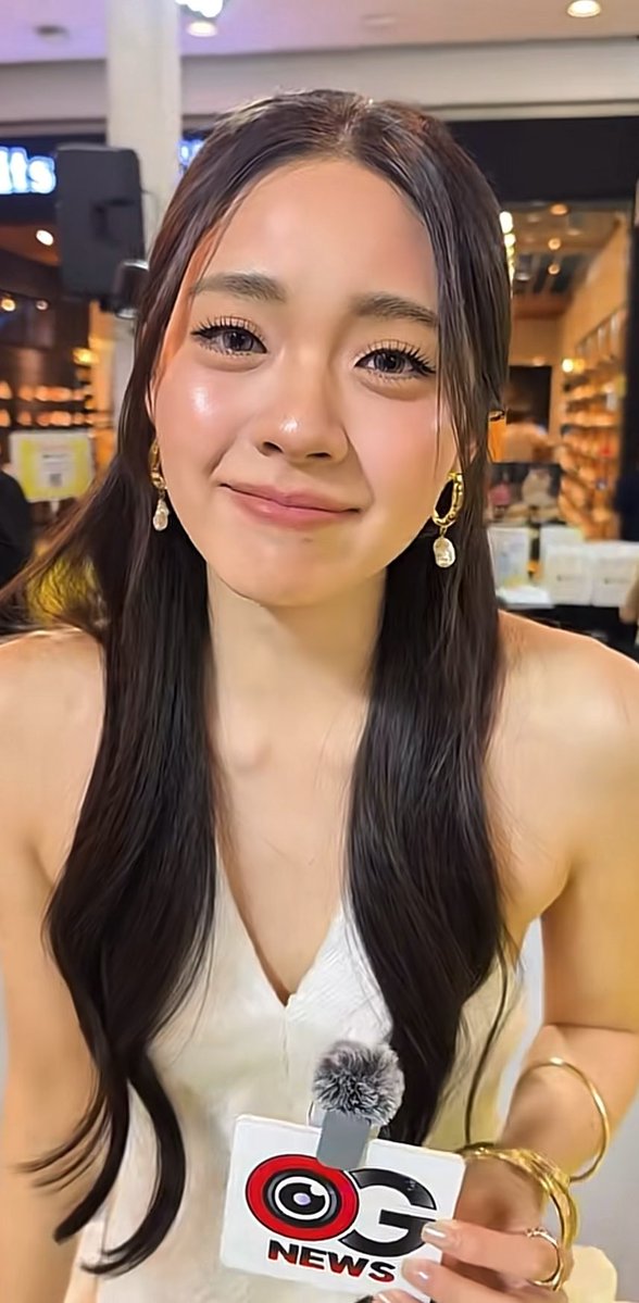 A young woman with long dark hair wearing a white top and dangling earrings smiles at the camera while holding a CG News microphone with a furry windscreen.