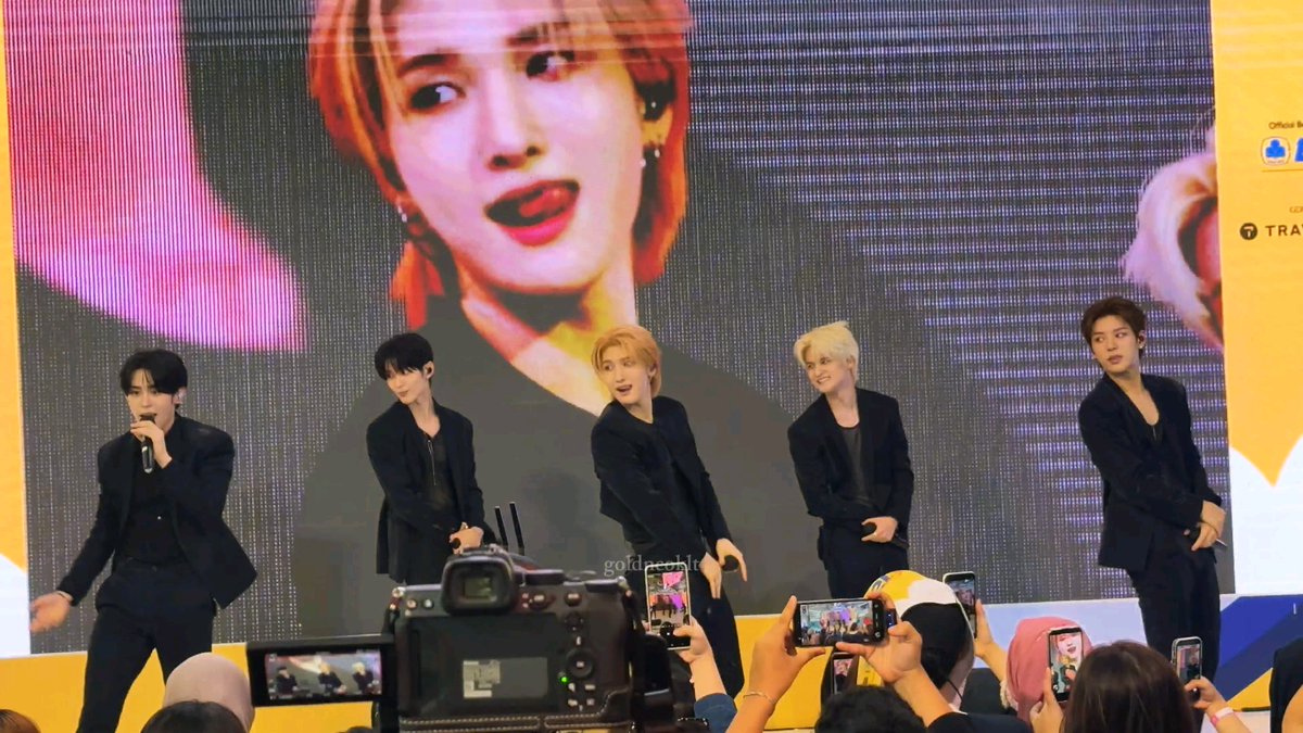 A five-member K-pop boy group performs on stage in black suits in front of a large screen displaying a woman with red hair, while audience members hold up phones to record, watermarked "gold.pebbl".