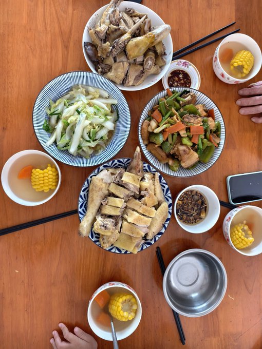 A family meal on a wooden table featuring plates of chicken, bok choy, stir-fried vegetables with mushrooms, bowls of corn soup, and condiments.