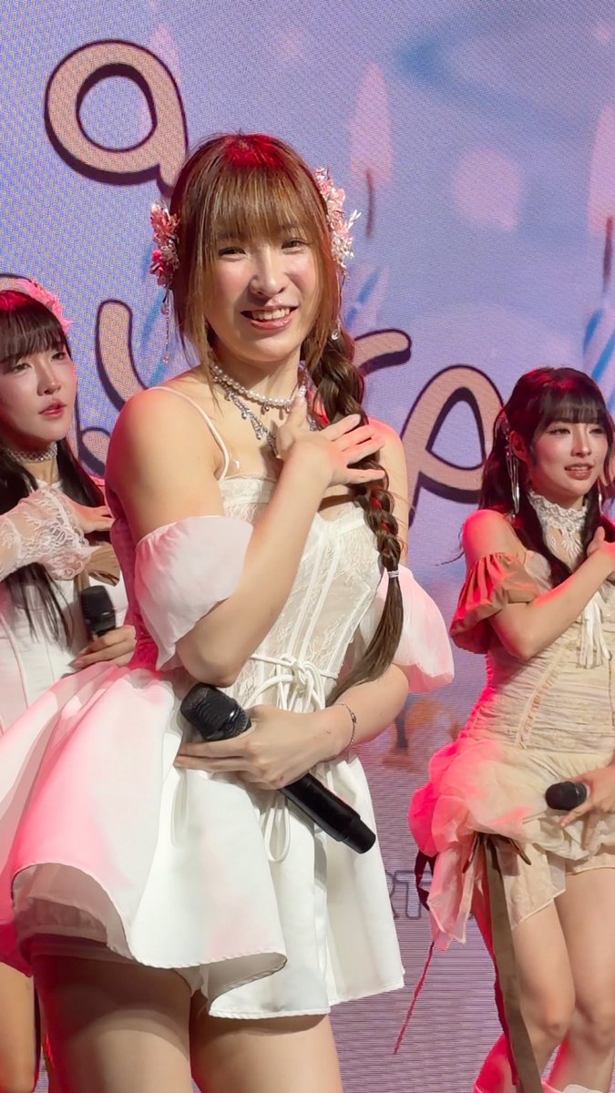 A smiling woman with brown hair and floral hair accessories performs on stage in a white corset dress, holding a microphone alongside other performers in pink outfits.
