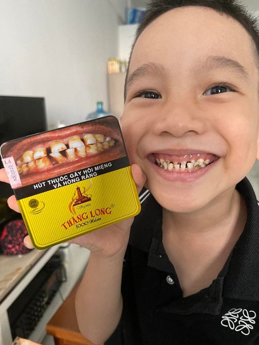 A young boy smiling and holding up a yellow tin of 'Thang Long' betel nut (Hút thuốc gây hại miệng và hông răng) with a grim image of damaged teeth on the packaging.