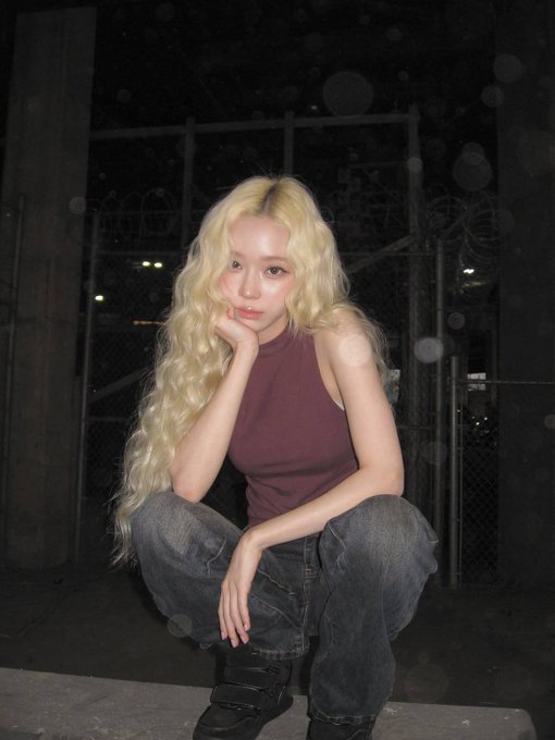 A nighttime portrait of a young woman with long, wavy blonde hair wearing a maroon halter top and baggy jeans, squatting on a sidewalk.