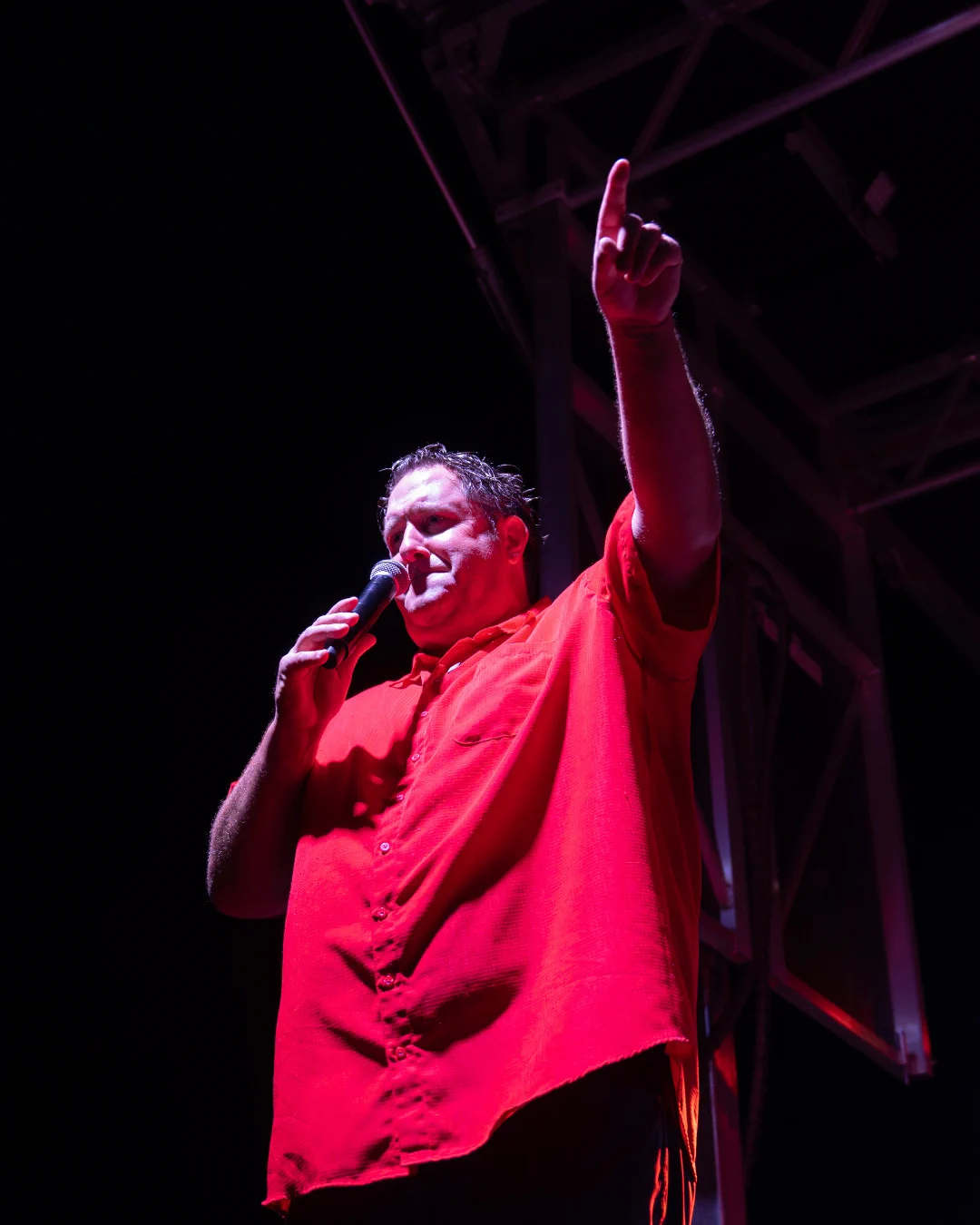 Coach Eck wearing a bright red shirt is speaking into a microphone on stage, under strong lighting.