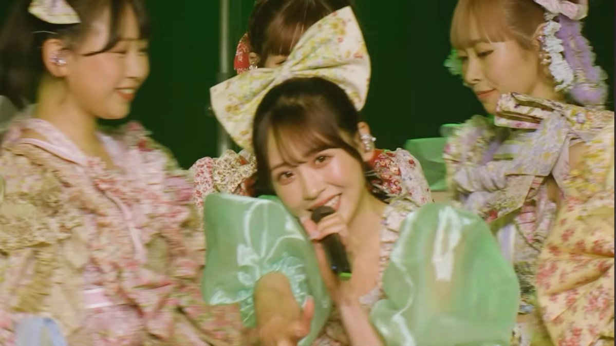 A group of performers in matching floral costumes with large bows, including one holding a microphone and leaning forward with a playful expression, perform on stage. The image corresponds to the post about Yui-chan (Sakurai Yui), likely showing her center stage in a cheerful, candid moment during a live performance.
