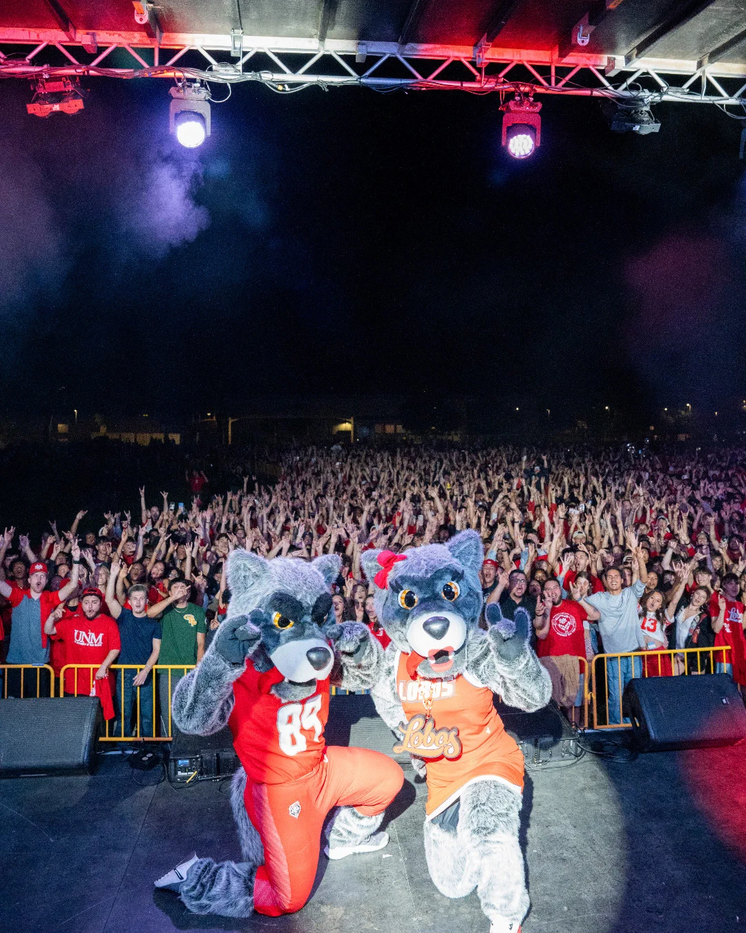 Louie and Lucy in red University of New Mexico Lobos uniforms pose on stage in front of a large, enthusiastic crowd. The audience in the background appears excited, with many raising their hands. Stage lights illuminate the scene.