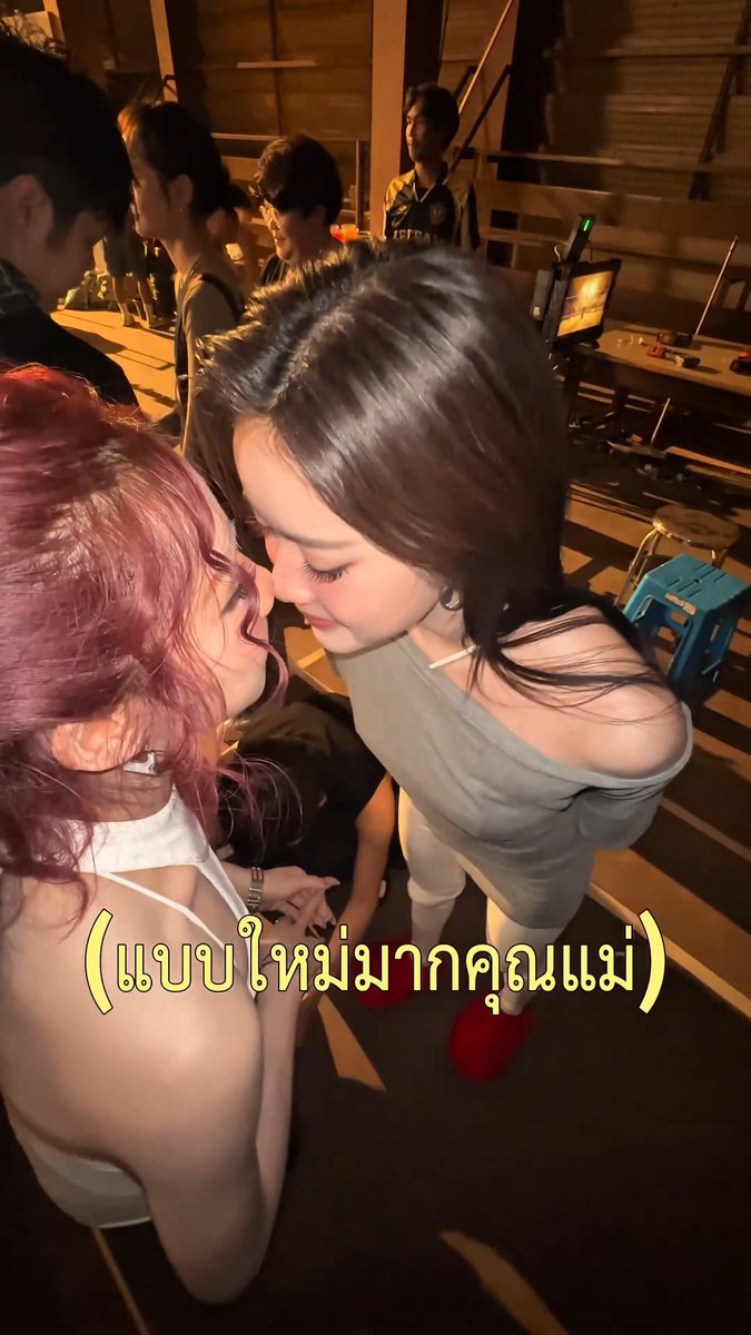 Two young women pose playfully close together at a fan event or gaming venue, with Thai text '(แบบไหนมากคุณแม่)' overlaid on the image. One has pink curly hair and the other has long dark hair wearing a gray off-shoulder top.