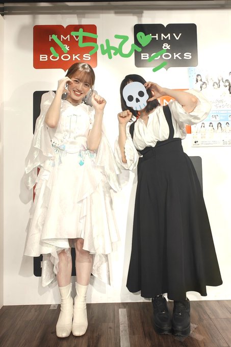 Two women pose in front of an 'HMV & BOOKS' backdrop with Japanese text 'ずっとそばに' (always by your side); one wears a flowing white dress and the other wears a black skirt with white blouse, holding a skull mask.
