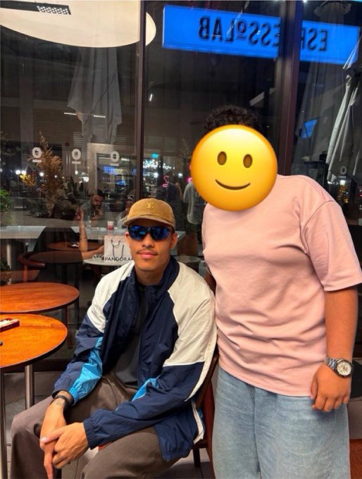 A Fenerbahçe fan, identifiable by the team's logo on his cap, sits at a coffee shop next to a tall man in a pink shirt whose face is obscured by a smiley emoji. The reflection on the window behind them shows "ESPRESSO LAB," matching the location mentioned in the post where the fan reportedly met Mason Greenwood.