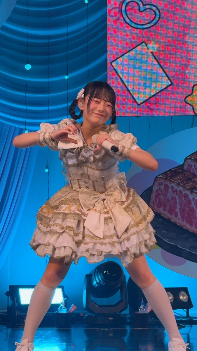 A performer on stage in a frilly cream-colored dress makes a heart shape with her hands while holding a microphone, smiling with her eyes closed.
