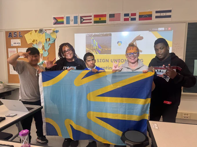 Group of three students with a teacher and staff member smiling at the camera while holding up the blue and yellow Sign Union flag, which represents the Deaf culture, community, heritage, and empowerment.