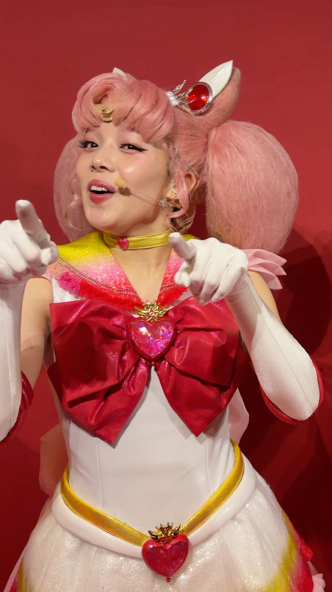 A performer dressed as Chibimoon (Sailor Chibi Moon) from Sailor Moon poses with a wide, slightly unsettling grin, wearing the character's signature pink pigtails, red bow, and white gloves while pointing forward. The caption references her "Shine on you" gesture and "creepy laugh," both of which are captured in this animated, mid-laugh moment of the costumed character.