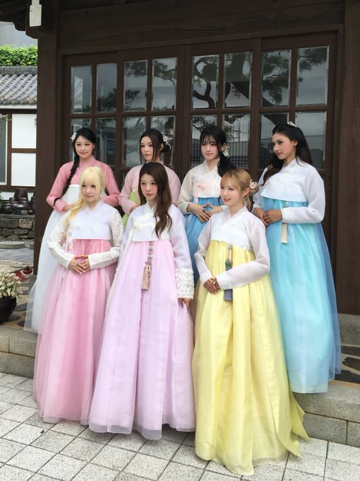 A group of seven young women pose together in colorful traditional Korean hanbok dresses in pastel shades of pink, blue, yellow, and white in front of a traditional building.