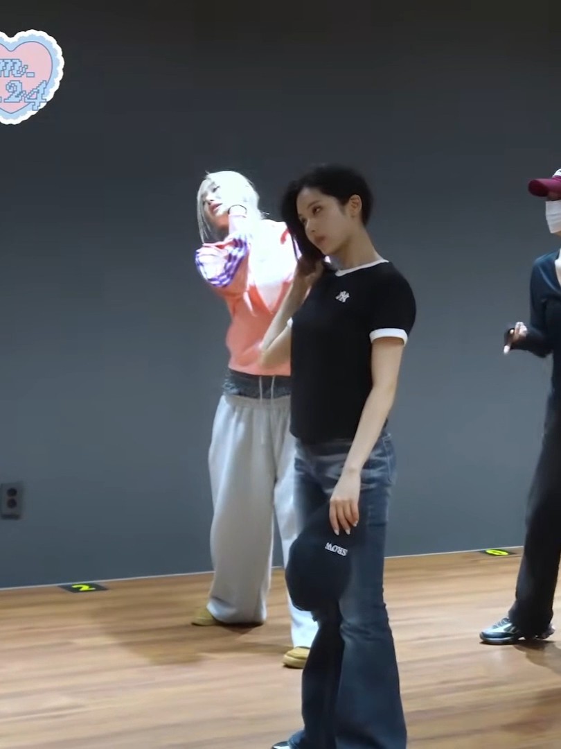 Two women are in a dance practice room, one in the foreground wearing a black t-shirt and jeans, while another with blonde hair dances in the background wearing an orange and white top.
