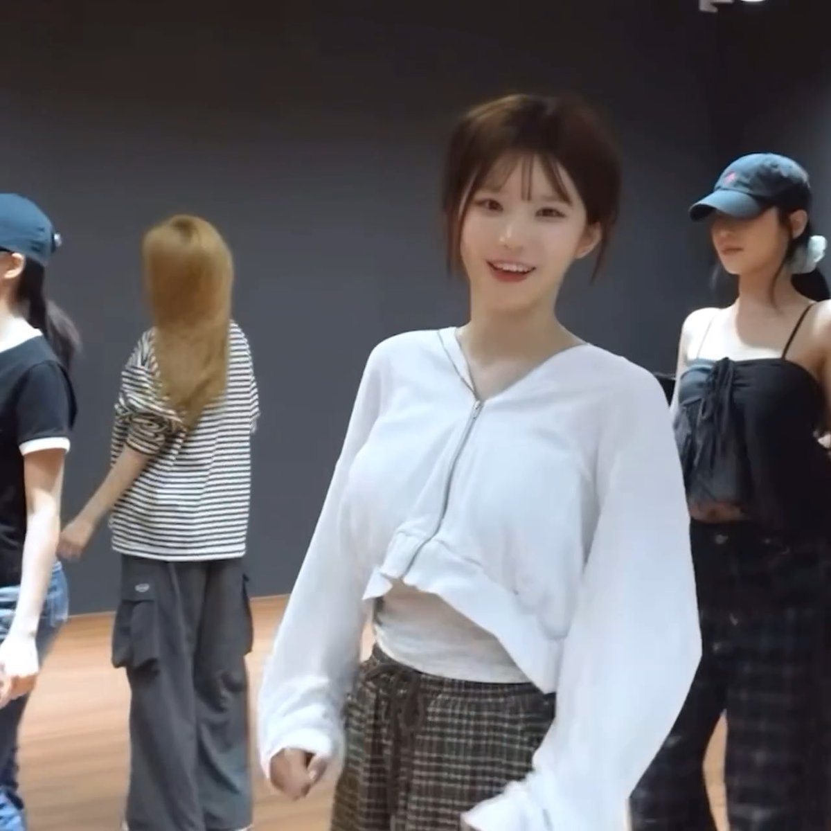 A young woman with short brown hair smiles at the camera in what appears to be a dance studio, wearing an oversized white zip-up hoodie and plaid pants, with other dancers in casual outfits visible in the background.