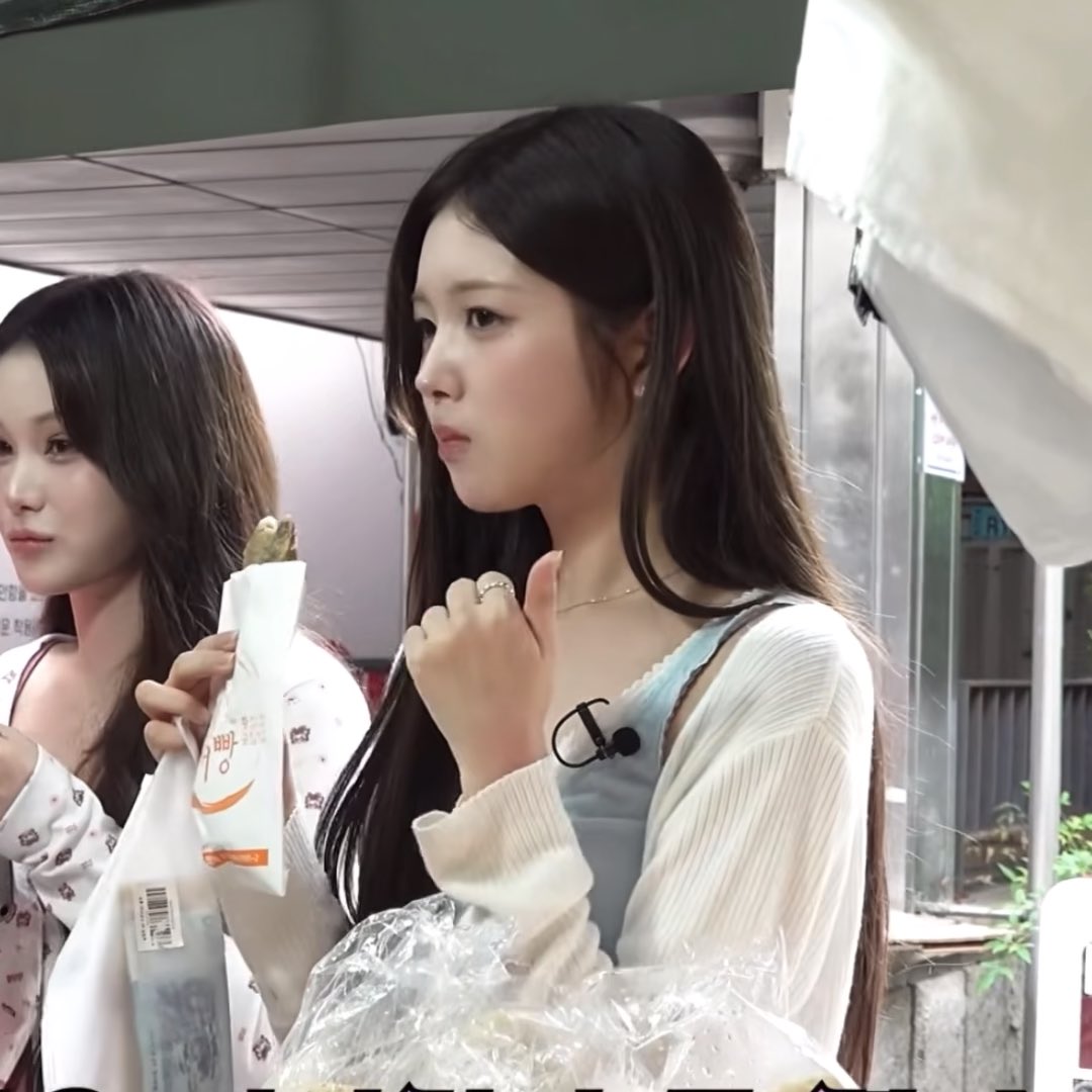 Two young women standing outdoors, with the one in the foreground wearing a light blue top and white cardigan while holding a paper-wrapped food item and giving a thumbs up.