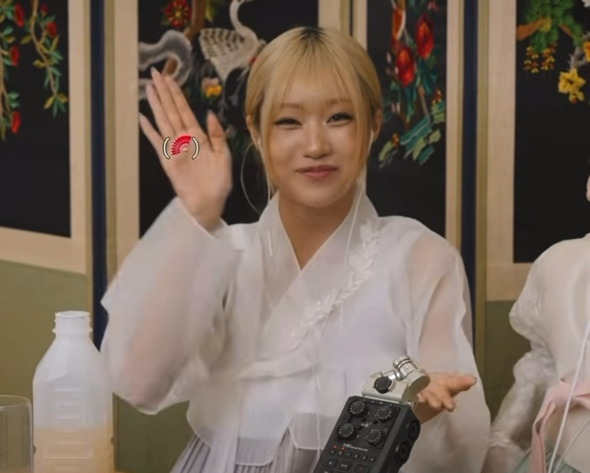 A young woman with blonde bangs wearing a white traditional Korean hanbok top waves cheerfully while holding an audio recorder, seated in front of decorative panels.