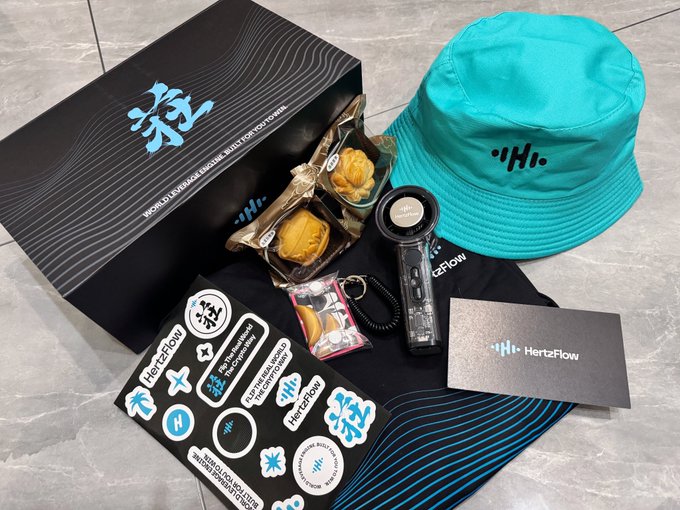 A HertzFlow promotional package displayed on a marble surface, featuring a teal bucket hat, a black box with "WORLD LIVING ROOM & MUSIC FOR EVERYONE," a sticker sheet, a business card, a USB device, mooncakes, and snacks.