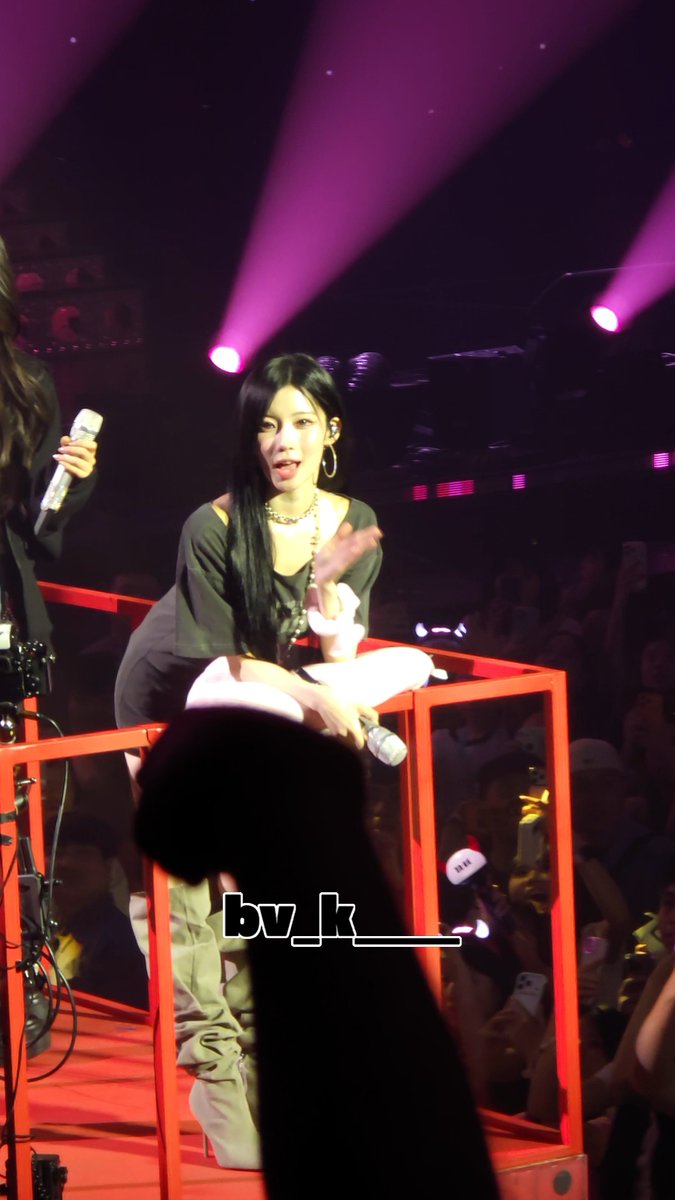 A performer with long dark hair sits on a red elevated platform during a concert under pink stage lights, holding a microphone and wearing a sheer top over a black bra, with the watermark 'bv_k___' visible.
