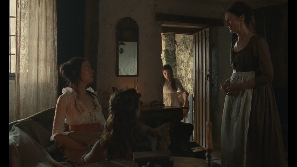 A dimly lit, cinematic scene from a period film shows four women in 18th-century attire gathered in a rustic room, with one standing in a doorway smiling at others seated near a window.
