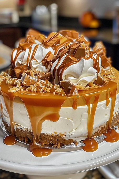A decadent caramel-drizzled cheesecake topped with whipped cream rosettes and chocolate candy pieces on a white cake stand.