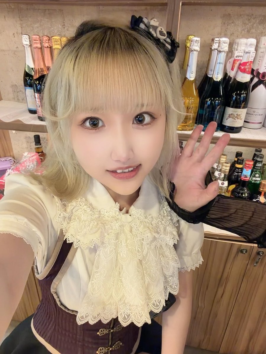 A selfie of a person with blonde hair wearing a Gothic Lolita style outfit with lace details and a panda hair clip, posing in front of a shelf filled with wine and champagne bottles.