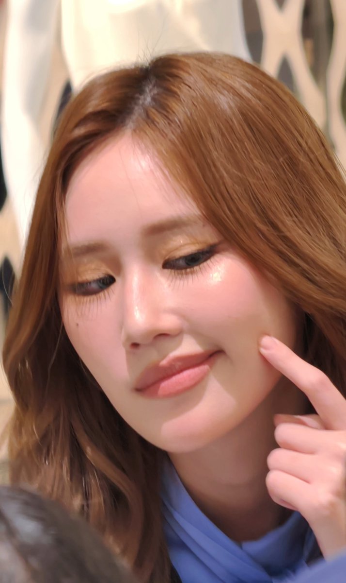 A close-up selfie of a young woman with reddish-brown hair, glossy makeup, and wearing a blue top, pointing a finger toward her cheek.