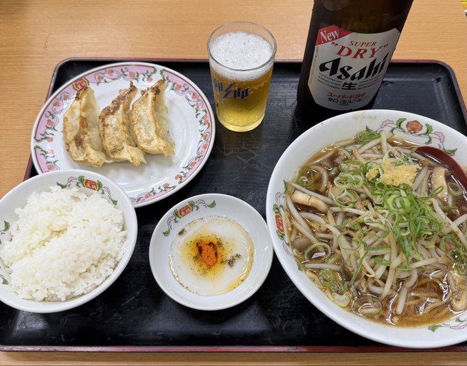 A Japanese meal on a tray featuring gyoza, a bowl of rice, a small side dish, a bowl of noodle soup, a glass of beer, and a bottle of Asahi Super Dry beer.
