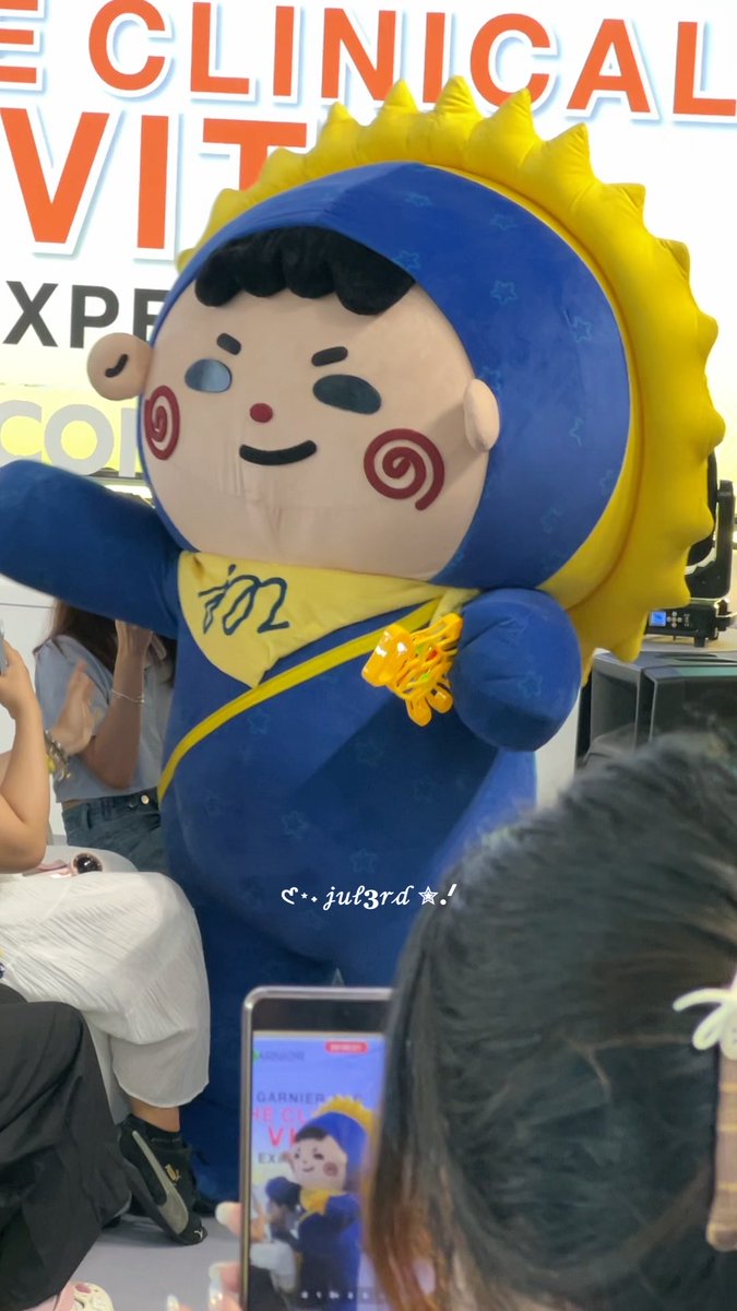 A large mascot character with a blue hood, yellow sun-like crown, and red swirl cheeks stands in front of a 'CLINICAL V' sign, while people in the foreground take photos.