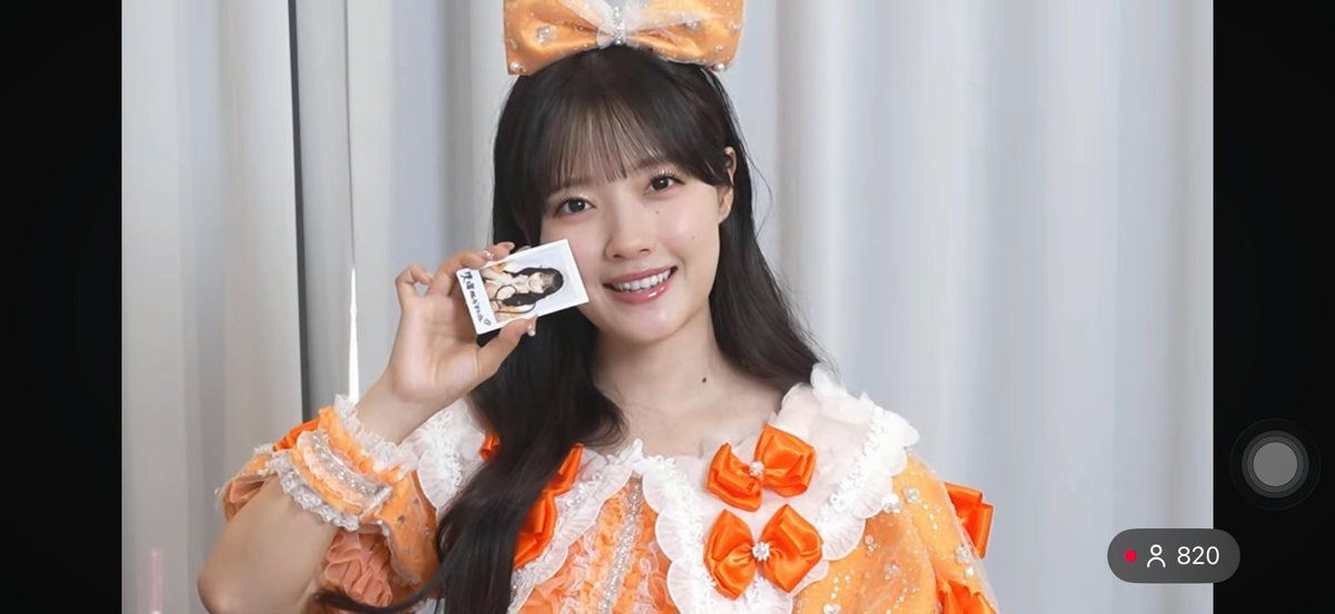 A young woman wearing an orange and white frilly idol outfit with a large orange bow is smiling and holding up a small photo card, with a viewer count of 820 visible on the right.