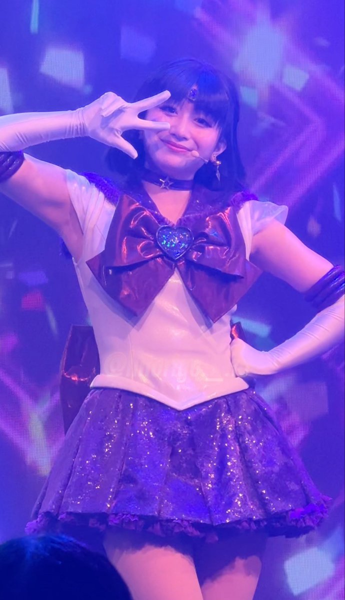 A performer in a Sailor Moon-inspired cosplay costume (white and purple with a large burgundy bow) wearing a headset microphone and white gloves, posing with a peace sign on a stage with purple lighting.