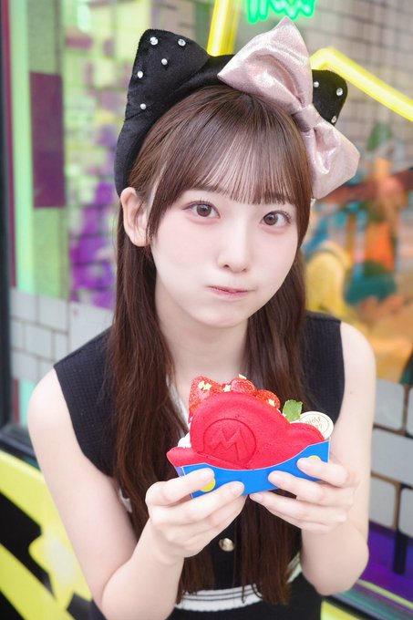 A young woman wearing a black top and decorative bow headband holds a large, colorful dessert shaped like a Mario mushroom.