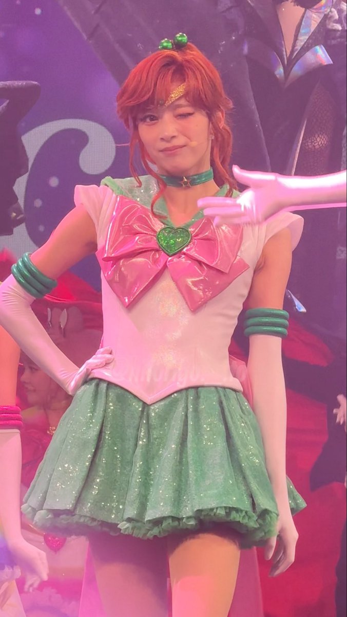 A performer in a Sailor Jupiter-inspired costume with red hair, a green and pink outfit featuring a heart-shaped brooch, winks at the camera during a stage performance.
