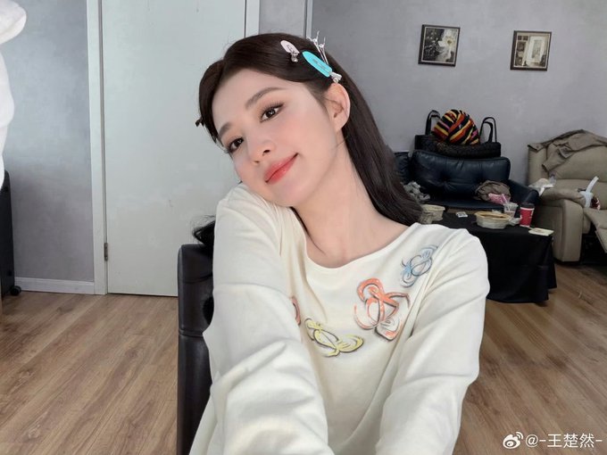 A selfie of a smiling woman with hair clips wearing a cream-colored top with colorful embroidered details, sitting indoors with a watermark reading '@王楚然-' at the bottom.