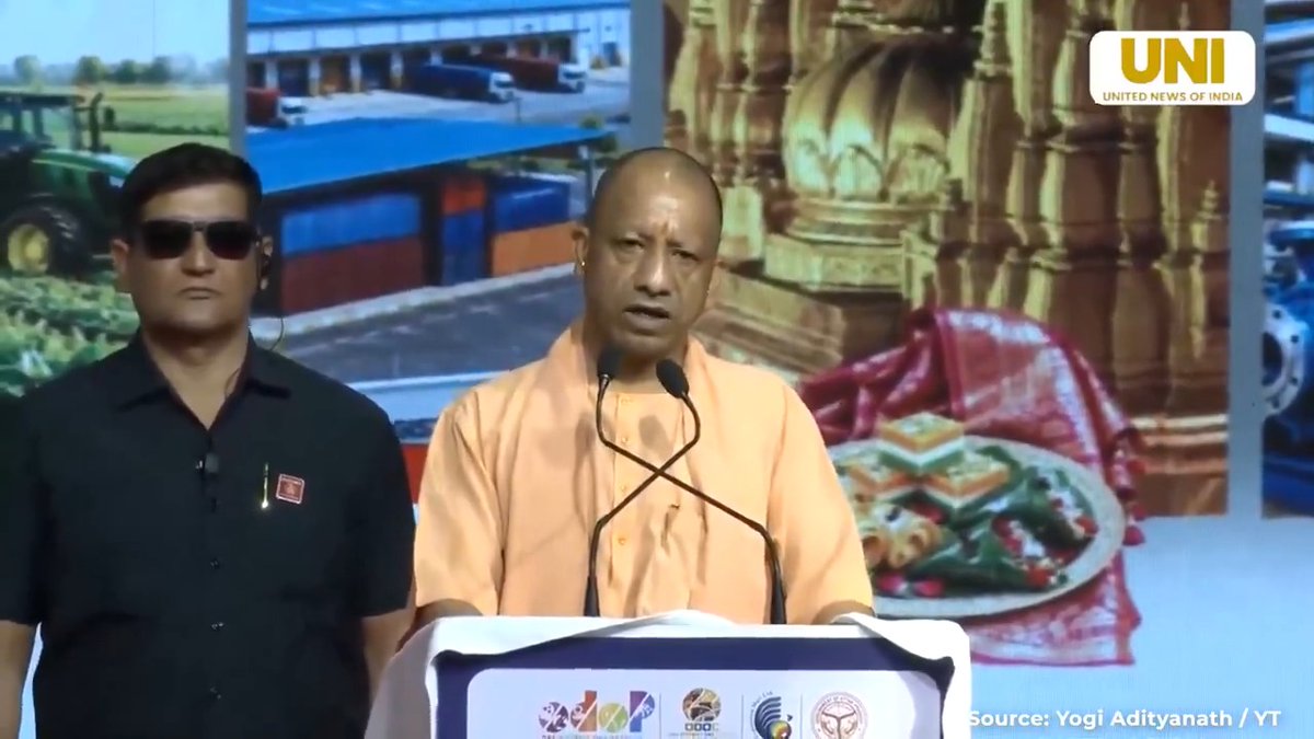 Yogi Adityanath, Chief Minister of Uttar Pradesh, speaks at a podium wearing his signature saffron robes, with a security officer in sunglasses standing beside him; the background shows images related to Uttar Pradesh development, and the source is credited as 'Yogi Adityanath / YT' via UNI (United News of India).