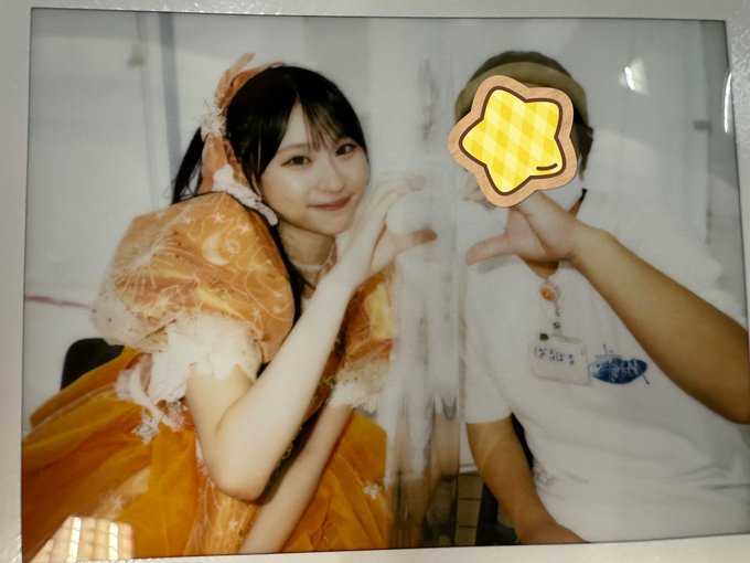 A young woman in an orange frilly dress poses with another person whose face is obscured by a yellow star sticker, both holding up finger hearts.