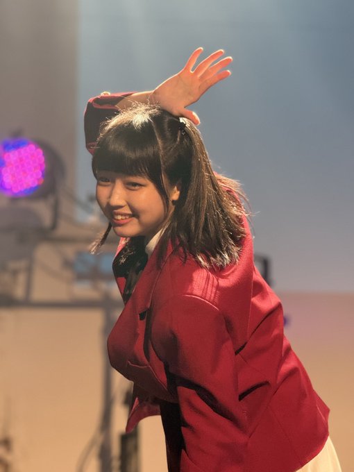 A young woman in a red blazer smiling and waving, with her dark hair in pigtails, on what appears to be a stage with lights in the background.