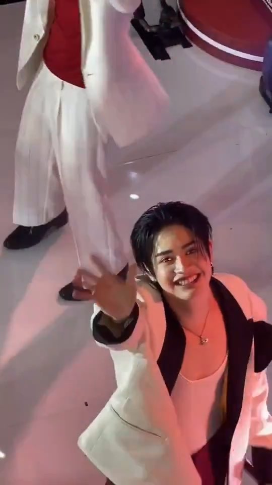 A smiling person with dark hair wearing a cream blazer with black lapels and a pink top gestures enthusiastically toward the camera, with another person in white partially visible behind them.