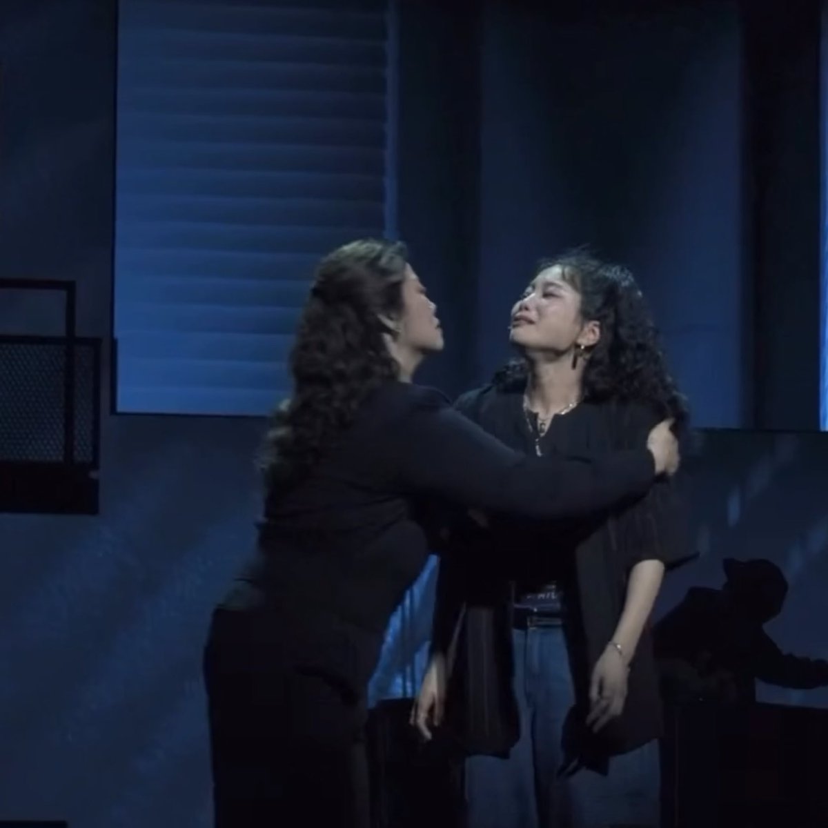 Two women with curly hair embrace on a dimly lit stage with blue lighting, appearing emotional during a theatrical performance.