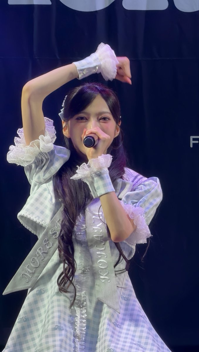 A young woman on stage sings into a microphone, wearing a light blue and white checkered dress with lace details and sashes reading "MORE Sx" and "LINKCLICK".