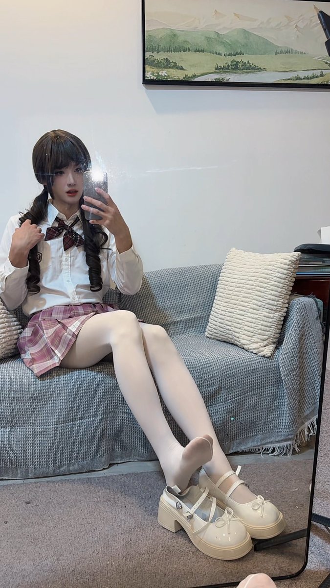 A young woman takes a mirror selfie on a gray couch, wearing a school uniform-style outfit with a white shirt, plaid skirt, and white tights, paired with chunky cream Mary Jane shoes.