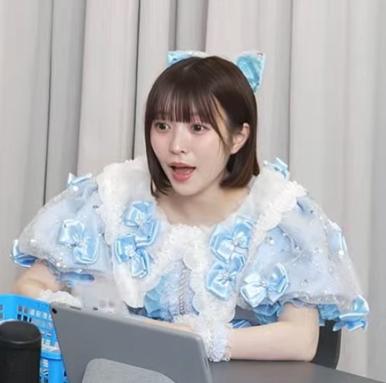 A woman with short brown hair and a blue bow headband, dressed in an ornate light blue and white ruffled outfit, speaks while looking at a laptop.