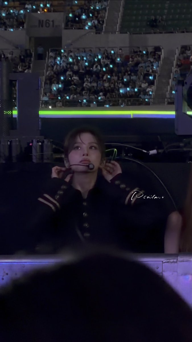 A young person wearing a headset with a microphone at what appears to be a concert or event, looking upward with stadium seating full of fans holding light sticks in the background. Watermark reads 'Pcostar'.