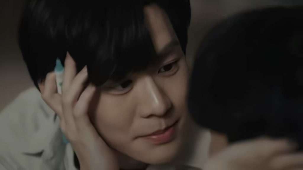 A young man with dark hair gently holds the head of another person, gazing at them with a soft smile.
